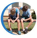 Two boys sitting on a bench, one looking sad and the other with his arm around him
