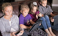 Children sitting on couch using various types of electronics or screens.