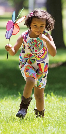 Photo of little girl wearing a sundress and holding a pinwheel running in a field of grass.